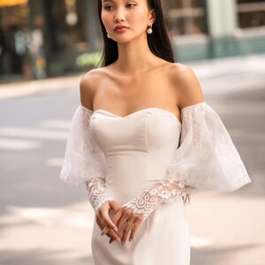 May include: A white off-the-shoulder bridal gown with a fitted bodice and a flowing skirt. The gown features delicate lace detailing on the sleeves and a sweetheart neckline.