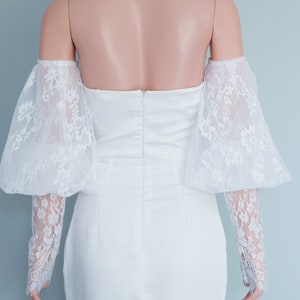 May include: White off-the-shoulder bridal dress with a fitted bodice and a lace overlay on the sleeves. The dress has a back zipper closure and a slit in the skirt.