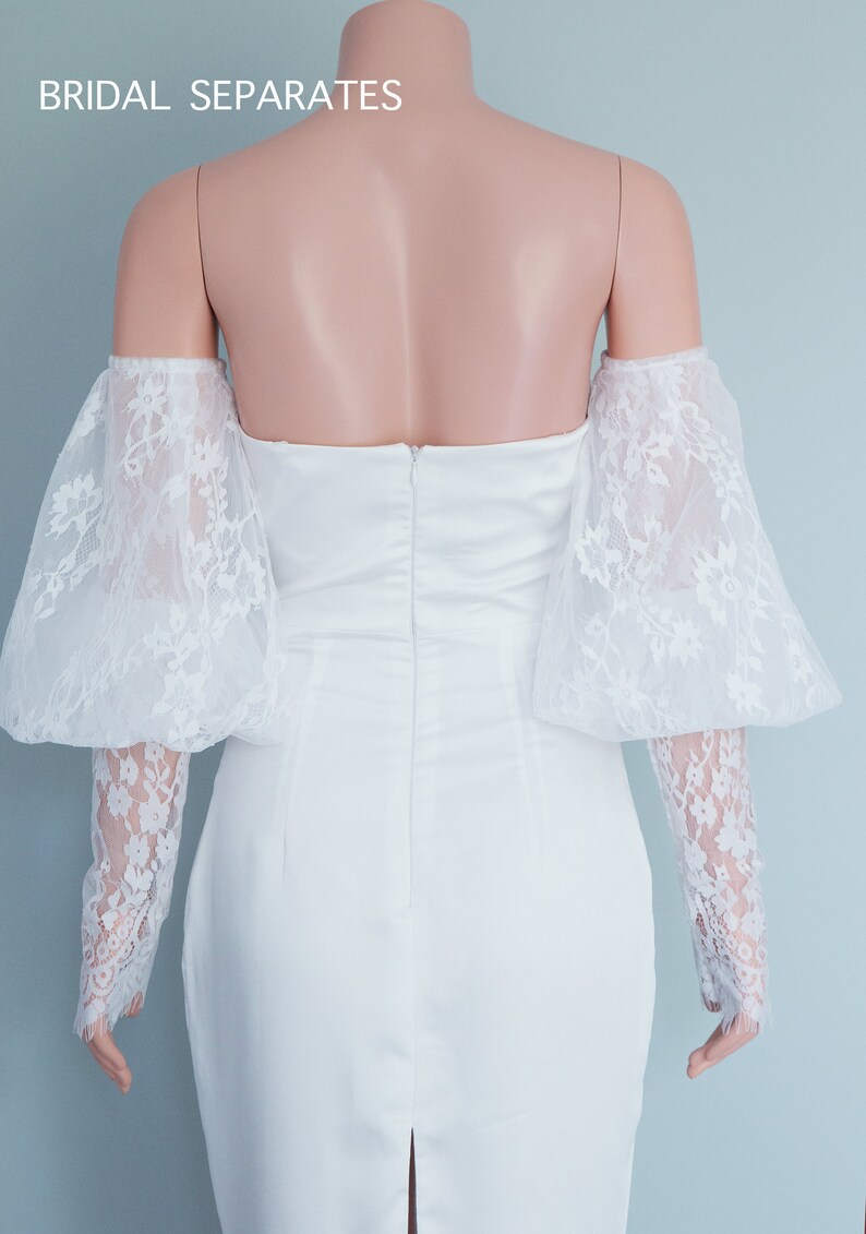 May include: White off-the-shoulder bridal dress with a fitted bodice and a lace overlay on the sleeves. The dress has a back zipper closure and a slit in the skirt.