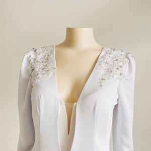 May include: A white long-sleeved dress with a deep V-neckline and beaded embellishments on the shoulders.