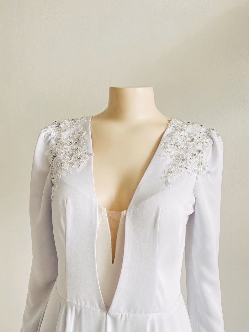 May include: A white long-sleeved dress with a deep V-neckline and beaded embellishments on the shoulders.