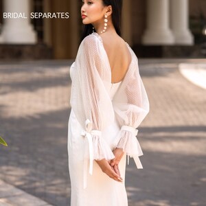 May include: A white bridal gown with a low back and sheer, polka dot sleeves. The sleeves have a ribbon tie at the wrist. The gown is photographed against a backdrop of a building with columns.