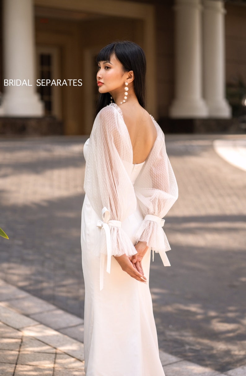 May include: A white bridal gown with a low back and sheer, polka dot sleeves. The sleeves have a ribbon tie at the wrist. The gown is photographed against a backdrop of a building with columns.