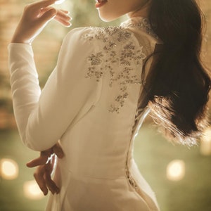 May include: A white dress with a long sleeve and a beaded design on the shoulder. The dress has a fitted bodice and a flowing skirt.