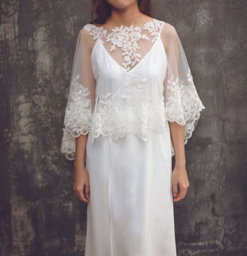 May include: A white lace overlay with a delicate floral pattern. The overlay is sheer and has a scalloped edge. It is worn over a white dress.