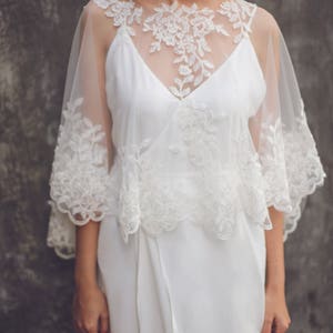 May include: A white lace overlay with a scalloped edge. The overlay is sheer and has a delicate floral pattern. It is worn over a white dress.