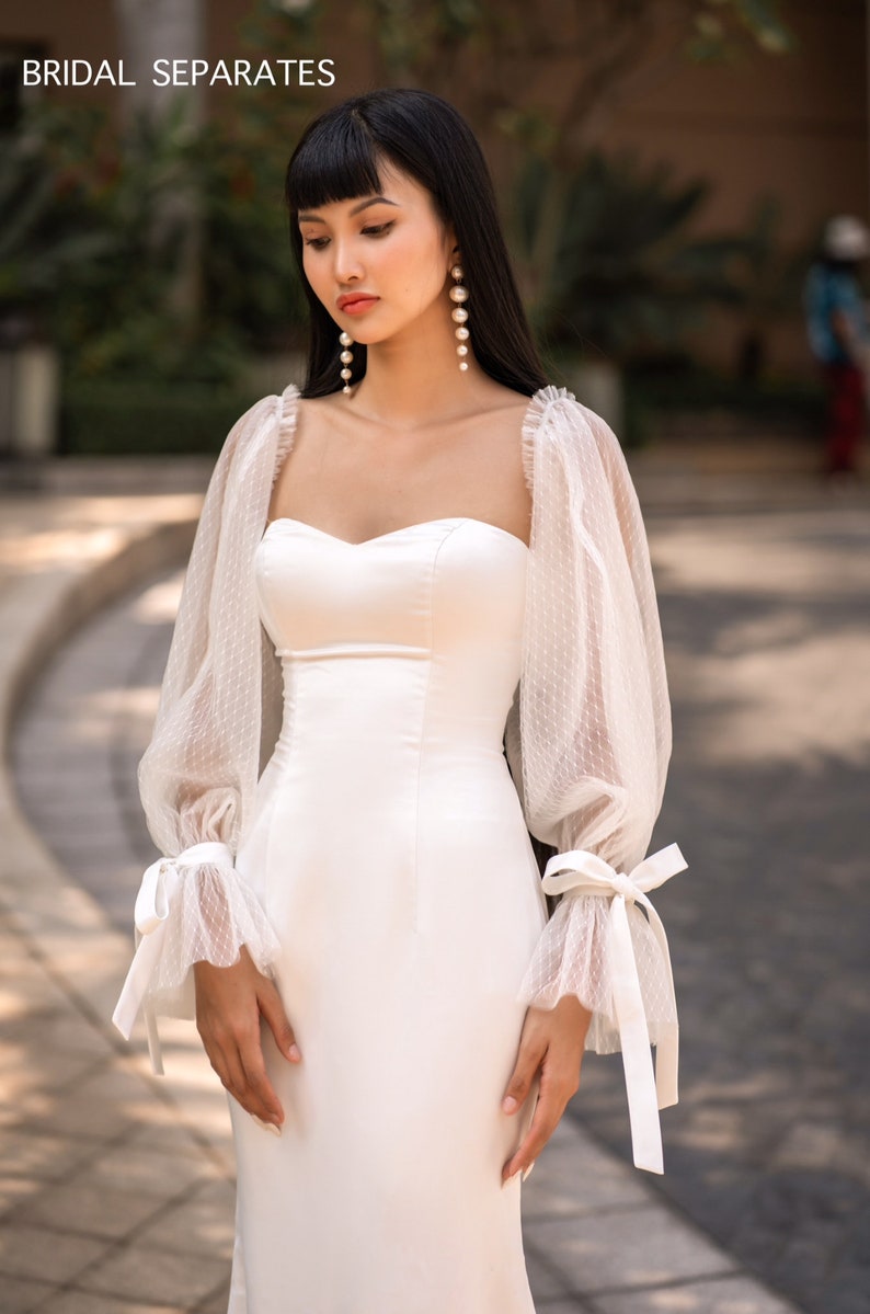 May include: A white bridal gown with a fitted bodice and a flowing skirt. The gown has long, sheer sleeves with delicate lace detailing and a bow at the wrist.