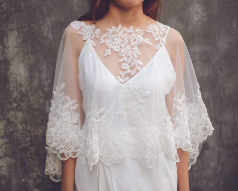 May include: A white lace bridal cape with a delicate floral pattern. The cape is sheer and has a scalloped edge.