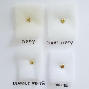 May include: Four swatches of fabric in ivory, light ivory, diamond white, and white. Each swatch has a small gold heart shape placed on it.