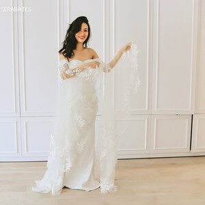 May include: A white off-the-shoulder wedding dress with lace detailing and a long veil. The dress is fitted and has a mermaid silhouette.