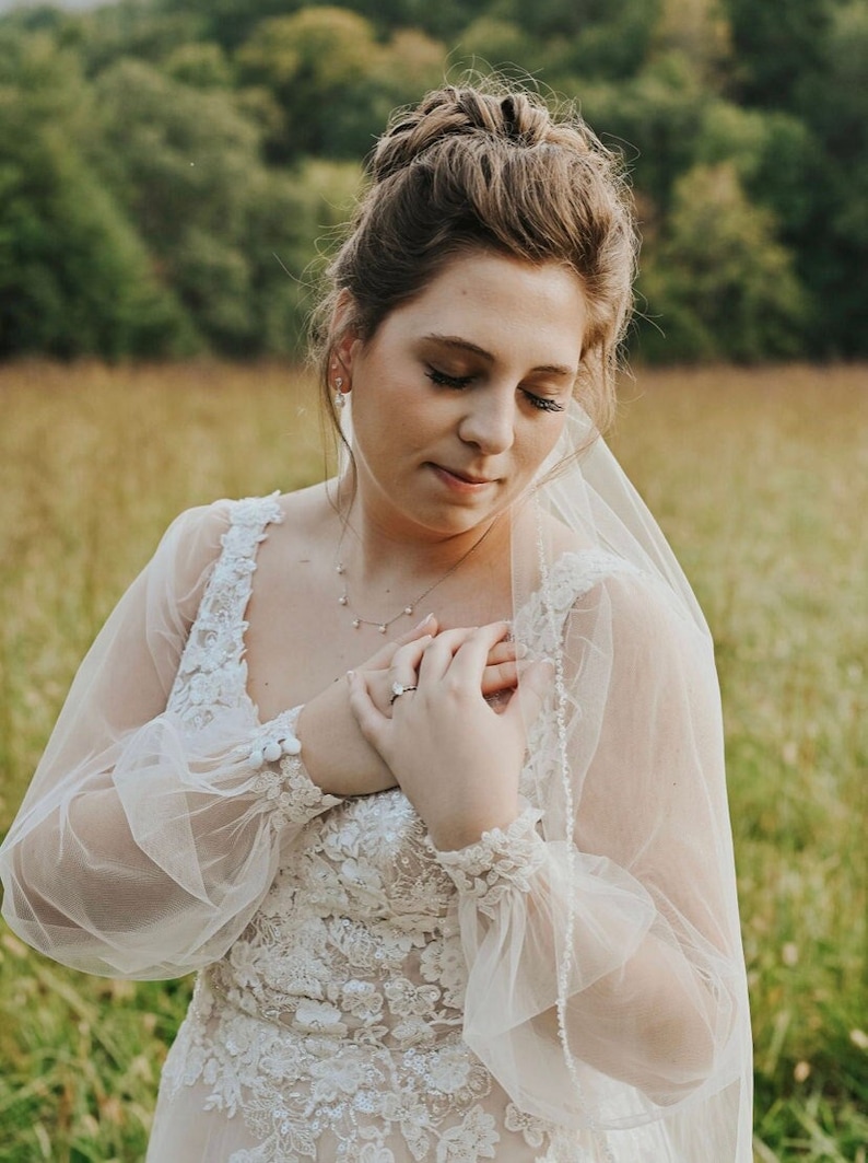 May include: A bride wearing a white lace wedding dress with a tulle veil. She is standing in a field of long grass and has her hands clasped over her heart.