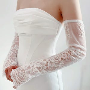 May include: White lace bridal gloves with a floral pattern. The gloves are made of a sheer fabric and have a delicate, feminine look.