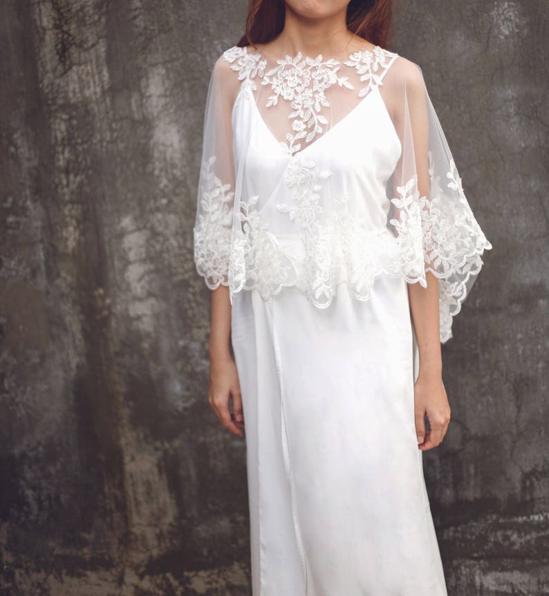 May include: A white lace bridal cape with a delicate floral pattern. The cape is sheer and has a scalloped edge. It is worn over a white dress.