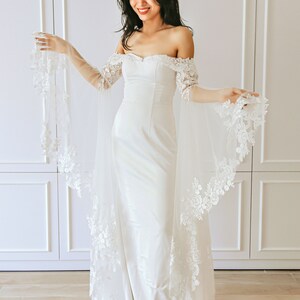 May include: A white off-the-shoulder bridal gown with a fitted bodice and a flowing skirt. The gown features delicate lace detailing on the sleeves and hem. The model is wearing the gown with a veil.