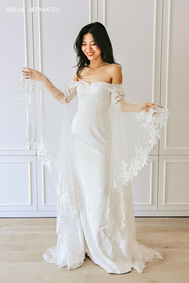 May include: A white off-the-shoulder bridal gown with a fitted bodice and a flowing skirt. The gown features delicate lace detailing on the sleeves and hem. The model is wearing the gown with a veil.