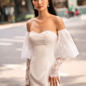 May include: A white off-the-shoulder bridal gown with a fitted bodice and a lace overlay on the sleeves. The gown has a sweetheart neckline and a long skirt.
