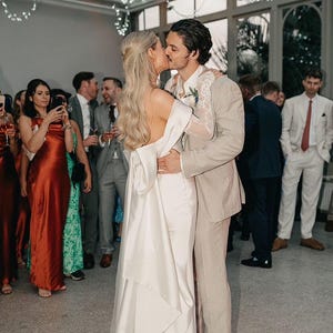 May include: A bride and groom are kissing during their wedding reception. The bride is wearing a white satin wedding dress with a long train. The groom is wearing a light brown suit. The couple is surrounded by guests.