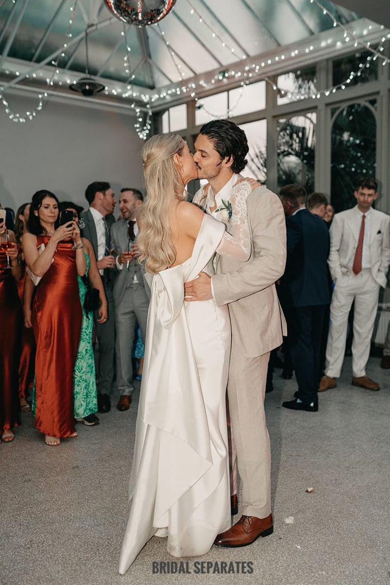 May include: A bride and groom are kissing during their wedding reception. The bride is wearing a white satin wedding dress with a long train. The groom is wearing a light brown suit. The couple is surrounded by guests.
