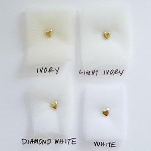May include: Four swatches of tulle fabric in different shades of white, labeled "Ivory", "Light Ivory", "Diamond White", and "White". A small gold heart is placed on each swatch.