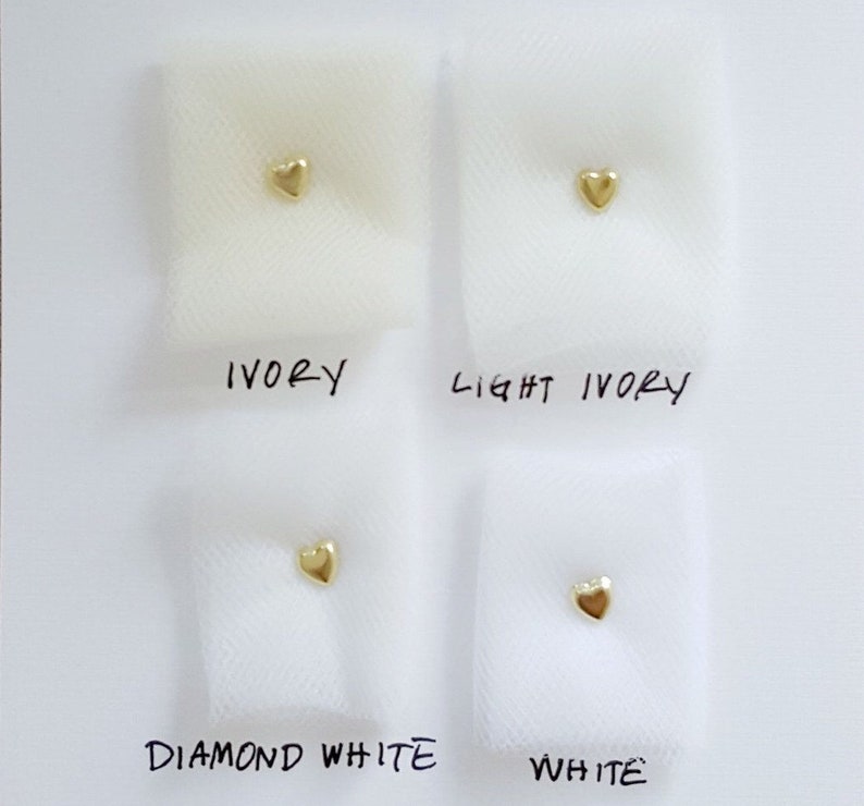 May include: Four swatches of tulle fabric in different shades of white, labeled "Ivory", "Light Ivory", "Diamond White", and "White". A small gold heart is placed on each swatch.