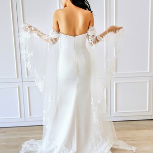 May include: A white bridal gown with a fitted bodice and a flowing skirt. The gown features off-the-shoulder straps and delicate lace detailing on the sleeves and train.