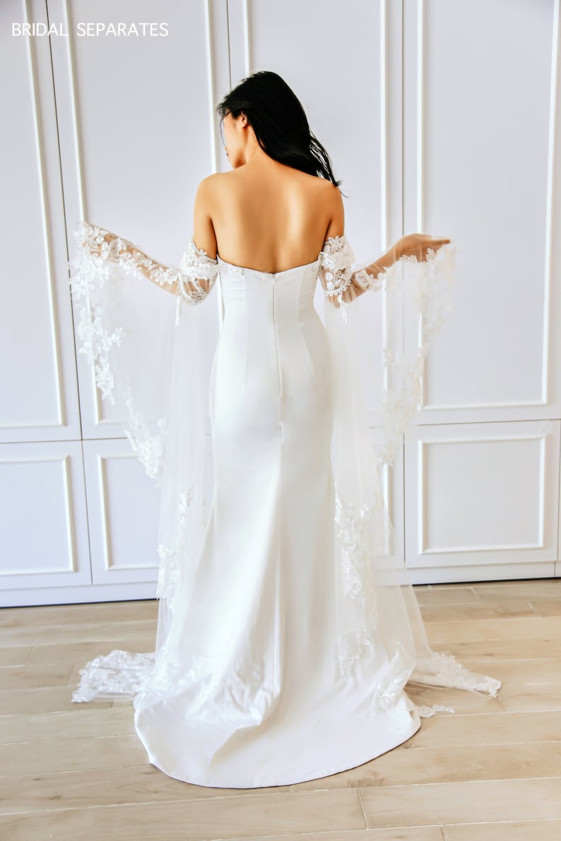 May include: A white bridal gown with a fitted bodice and a flowing skirt. The gown features off-the-shoulder straps and delicate lace detailing on the sleeves and train.