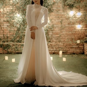 May include: A white wedding dress with a long train and a lace bodice. The dress has a long, flowing skirt and a fitted bodice with a high neckline. The dress is worn by a person standing in front of a brick wall with greenery.