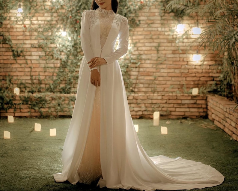 May include: A white wedding dress with a long train and a lace bodice. The dress has a long, flowing skirt and a fitted bodice with a high neckline. The dress is worn by a person standing in front of a brick wall with greenery.