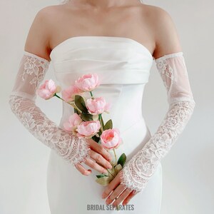 May include: White lace bridal gloves with a delicate floral pattern. The gloves are worn by a person holding a bouquet of pink flowers.