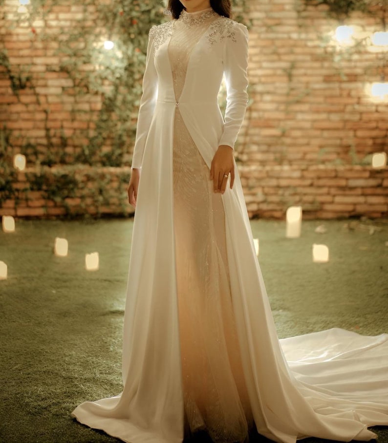 May include: A white bridal gown with a long train and a high neckline. The gown has a sheer overlay with sparkling embellishments and a fitted bodice.