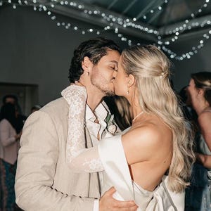 May include: A couple in a wedding setting, the bride is wearing a white satin dress with a large bow in the back, the groom is wearing a light brown suit jacket and a white shirt with lace sleeves. The couple is kissing.
