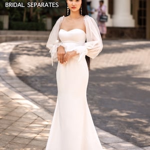 May include: A white bridal gown with a sweetheart neckline and long, sheer sleeves with puffed shoulders. The gown has a fitted bodice and a flowing skirt.