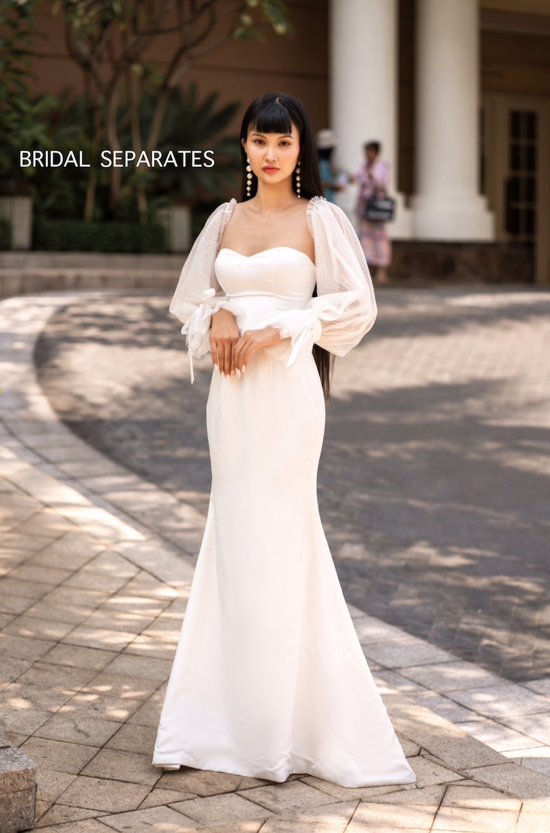 May include: A white bridal gown with a sweetheart neckline and long, sheer sleeves with puffed shoulders. The gown has a fitted bodice and a flowing skirt.