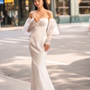 May include: A white off-the-shoulder bridal gown with a fitted bodice and a flowing skirt. The gown features delicate lace detailing on the sleeves and a simple, elegant design.