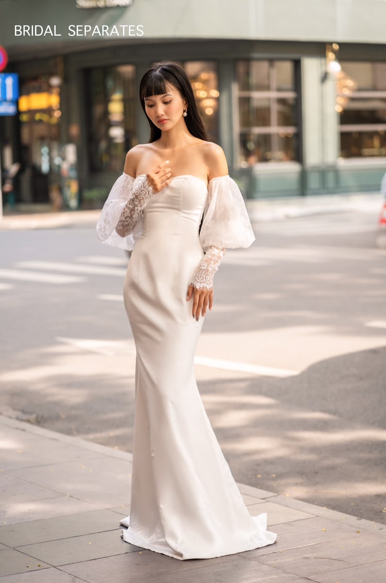 May include: A white off-the-shoulder bridal gown with a fitted bodice and a flowing skirt. The gown features delicate lace detailing on the sleeves and a simple, elegant design.