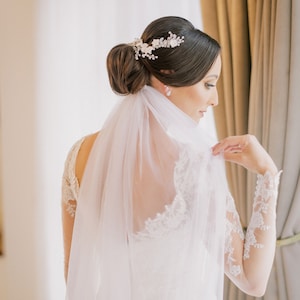 Pu&ograve; includere: Una sposa con un abito da sposa in pizzo bianco con un lungo velo. Ha i capelli raccolti in uno chignon con un accessorio per capelli floreale.
