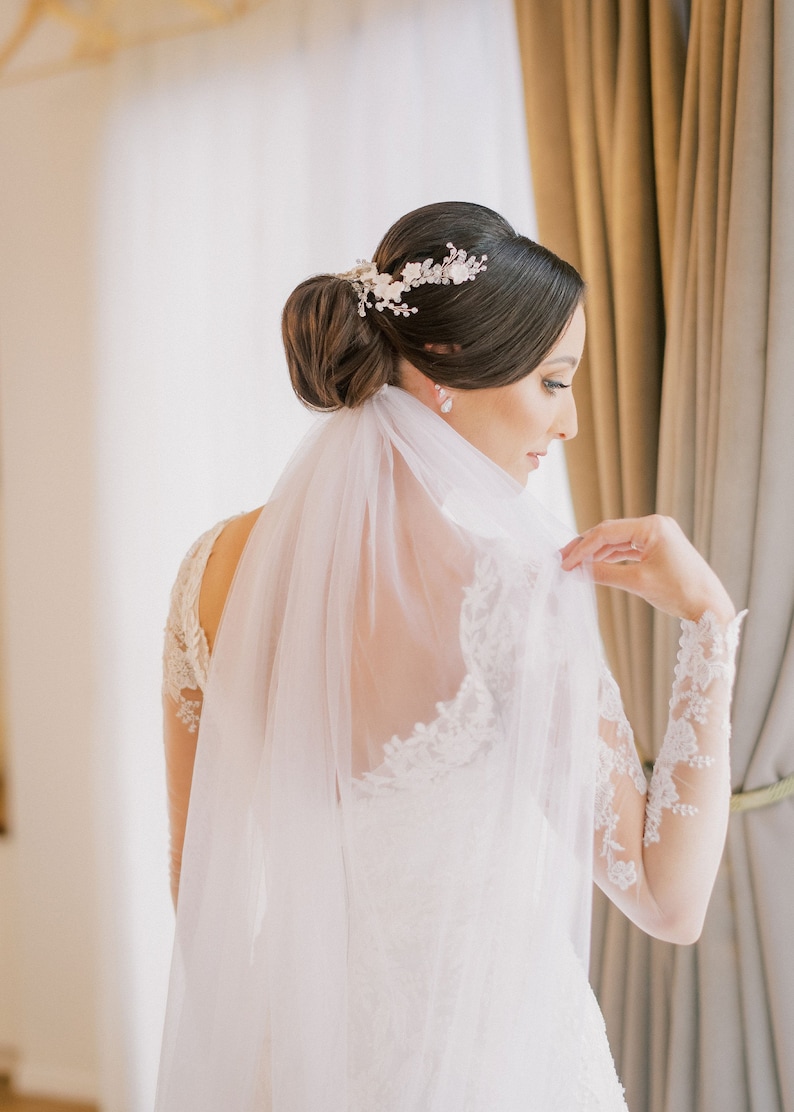 Pu&ograve; includere: Una sposa con un abito da sposa in pizzo bianco con un lungo velo. Ha i capelli raccolti in uno chignon con un accessorio per capelli floreale.