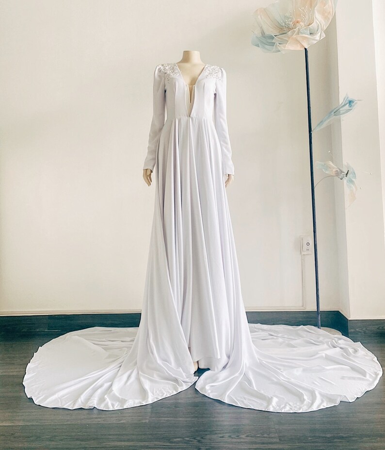 May include: A white long-sleeved gown with a plunging neckline and a flowing skirt. The gown has a beaded detail at the neckline.