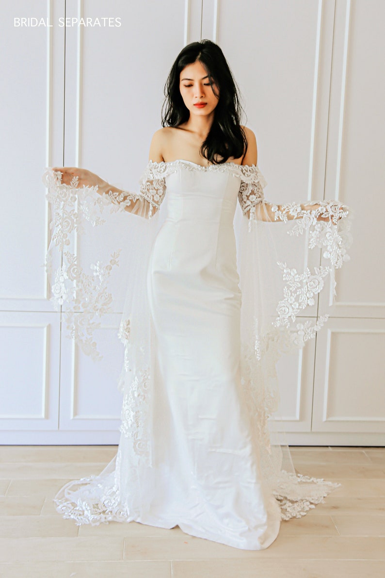 May include: A white off-the-shoulder wedding dress with a fitted bodice and a flowing skirt. The dress features delicate lace detailing on the sleeves and hem.