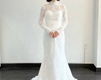 Puede incluir: Un vestido de novia de encaje blanco con cuello alto y mangas largas. El vestido tiene un corpiño ajustado y una falda fluida.