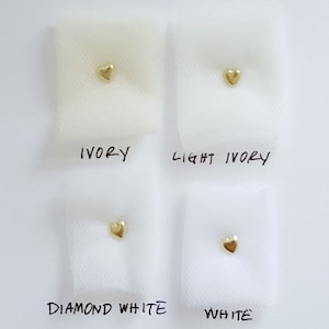 May include: Four swatches of tulle fabric in different shades of white, labelled "Ivory", "Light Ivory", "Diamond White", and "White". A small gold heart is placed on each swatch.