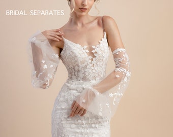 May include: A white bridal gown with a fitted bodice and a flowing skirt. The gown features delicate floral lace embroidery and sheer sleeves with floral accents. The model is wearing the gown with a simple hairstyle and minimal makeup.