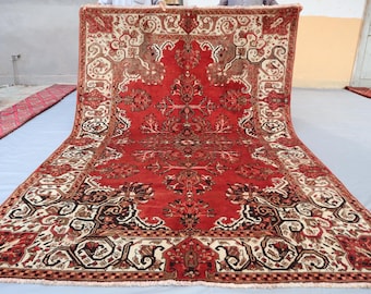 May include: A large, rectangular red rug with an intricate, symmetrical pattern. The design features floral and geometric motifs in shades of cream, brown, and black. The rug has a fringed edge and is displayed on a light blue surface.