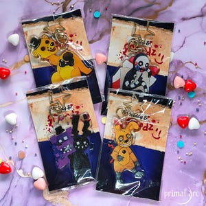 May include: Four colorful keychains featuring characters from the popular video game Five Nights at Freddy's. The keychains are made of clear acrylic and feature a variety of characters, including Freddy Fazbear, Chica the Chicken, Bonnie the Bunny, and Foxy the Pirate Fox. Each keychain has a gold metal ring for attaching to keys or bags.
