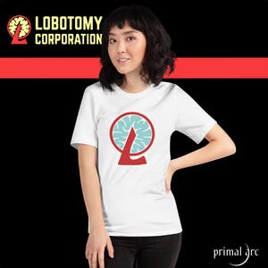 Può includere: Una maglietta bianca con un logo rosso e blu che presenta un cervello e il testo "Lobotomy Corporation".