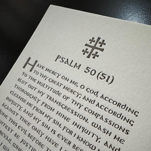 May include: Close-up of a textured, off-white paper with black text. The text includes the words "PSALM 50 (51)" and a religious symbol. The text is a passage from the Bible. The paper is slightly angled.