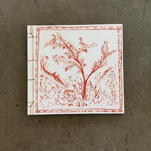 May include: A small, square, handmade book with a red and white illustration of birds perched on a tree. The cover features a detailed border and the pages are bound with a visible stitch.