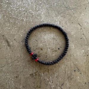 May include: A circular bracelet constructed from black knotted cord featuring a small cross shape formed by the knots, accented by two small, translucent red beads positioned on either side of the central cross element within the cord design.