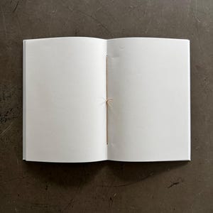 May include: An open, blank notebook with white pages and a tan string binding. The notebook is centered on a dark, textured surface. The pages are clean and ready for writing or sketching.