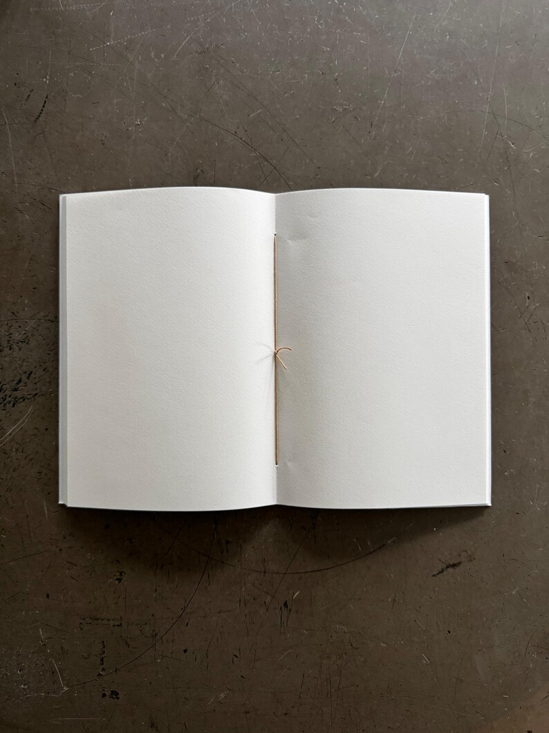 May include: An open, blank notebook with white pages and a tan string binding. The notebook is centered on a dark, textured surface. The pages are clean and ready for writing or sketching.